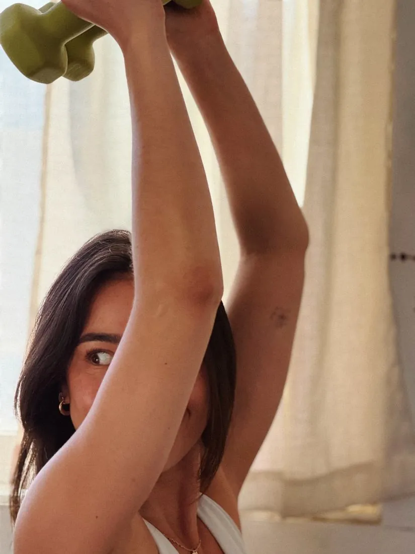 A woman lifting a green dumbbell overhead by a sunlit curtained window